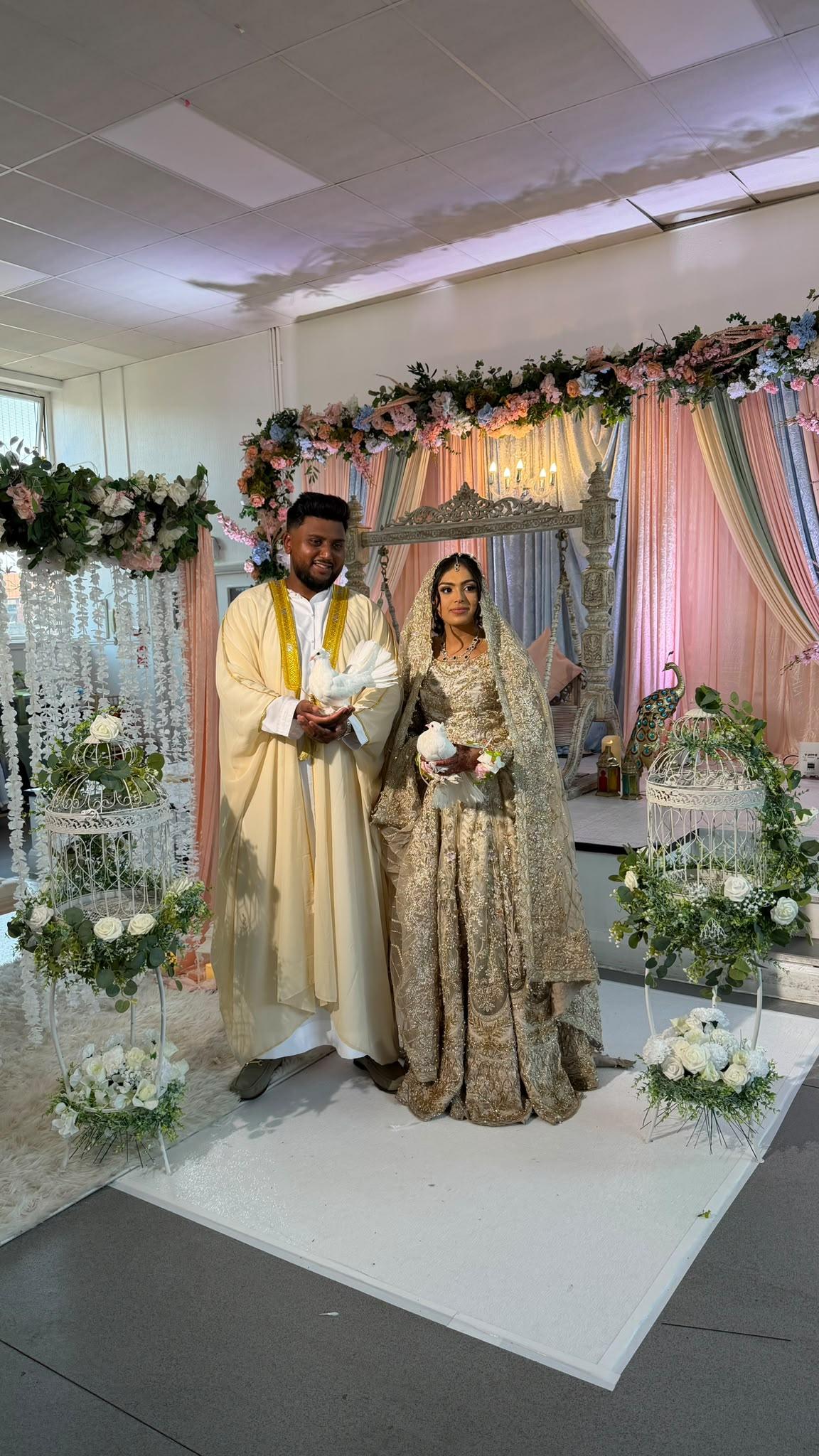 Bride and groom in traditional wedding attire each holding a white dove before a decorated stage